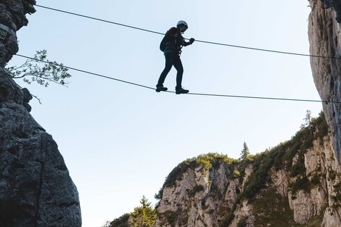 Eine Seilbrücke muss bei diesem Klettersteig von dem Sportler überquert werden. Er ist mit einem Klettersteigset gesichert.