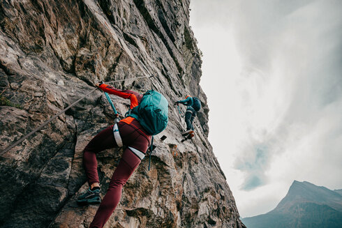 Eine Frau bewältigt gerade eine schwierige Passage am Klettersteig. Ein Mann schaut ihr von einer Stelle, die weiter oben liegt zu.